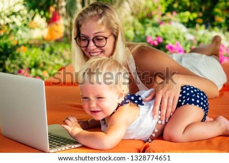 fair-haired one year old female kid looking at a laptop cartoon with mother blonde freelancer surfer on sunbeds resort summer beach.business woman freelancing and daughter relaxing by the sea