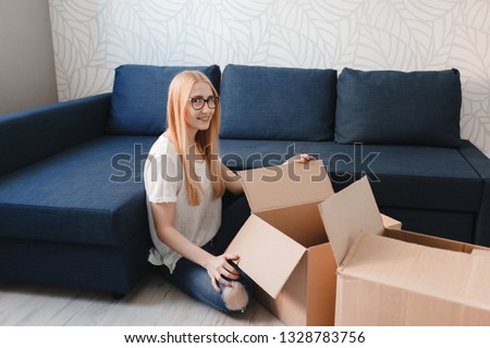 Moving house: Happy woman unpacking box in new home, blonde girl sittng near sofa with boxes