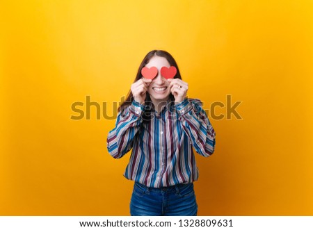 Lifestyle Concept: Attractive woman with beaming smile having two small red hearts in hands, closing eyes with paper heart symbols while standing over yellow background. 