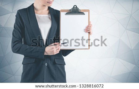 Portrait of unrecognizable young businesswoman wearing black suit and pointing at her clipboard with pen standing near geometric pattern wall. Mock up