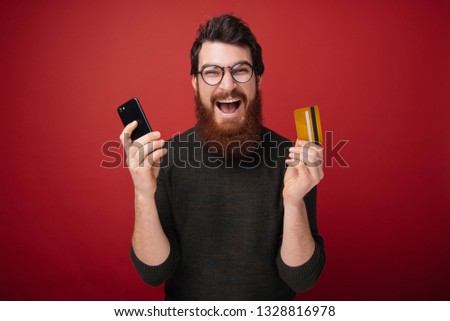 Photo of emotional excited bearded man standing isolated over red wall background holding credit card using mobile phone. Looking camera.