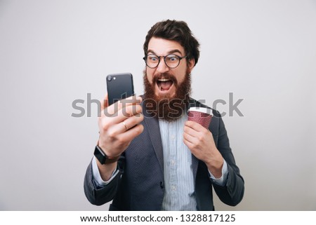 Bearded man wearing glasses using his mobile phone,with shocked face expression, holding takeaway coffee