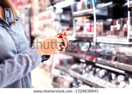 Woman buying make up at cosmetics section in store. Customer shopping beauty products. Choosing red lipstick from shelf and selection. Using tester. Makeup business concept. Supermarket or drugstore.