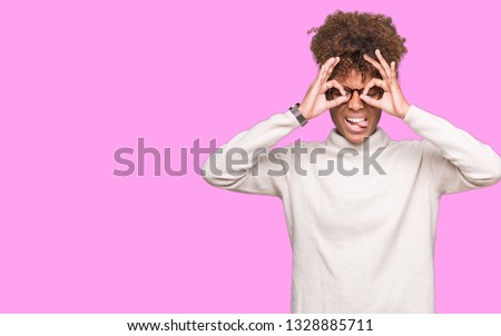 Beautiful young african american woman wearing glasses over isolated background doing ok gesture like binoculars sticking tongue out, eyes looking through fingers. Crazy expression.