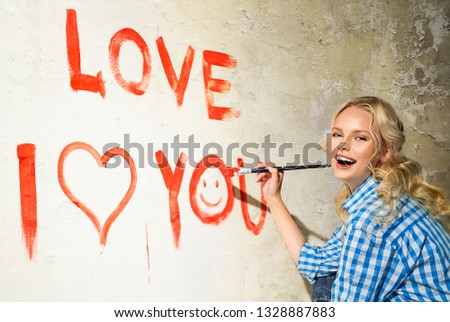 Young woman in plaid shirt with paintbrush is drawing a heart and inscription of love on the wall
