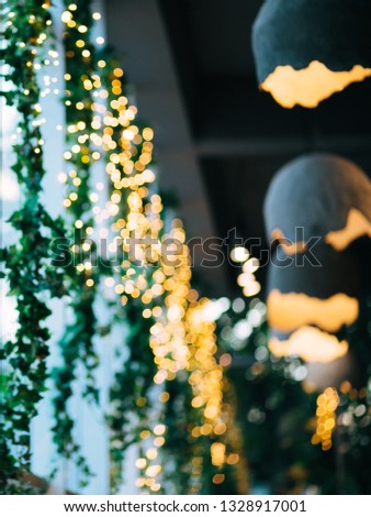 Lens blurred bokeh: multiple small bulbs, green leaves, chandeliers near a window. Indoor image.