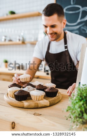 selective focus of barista standing behind bar counter and putting cupcakes on wooden tray in coffee house
