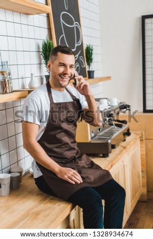 handsome cashier in brown apron talking on smartphone near coffee machine and sitting on bar counter in coffee house