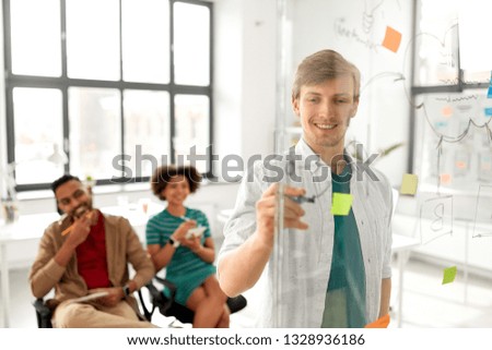 business, startup, planning and people concept - happy man drawing scheme on glass board for creative team at presentation in office