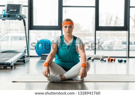 focused plus size girl sitting on fitness mat near sport equipment with crossed legs