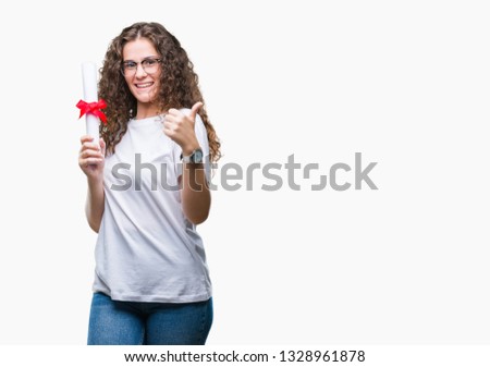Young brunette girl holding degree and wearing glasses over isolated background happy with big smile doing ok sign, thumb up with fingers, excellent sign