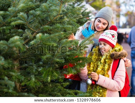 Happy satisfied pleasant smiling woman with daughter buying Christmas tree in market. Focus on woman