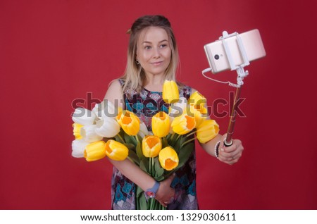 Beautiful woman making selfie on mobile phone with monopod holding bouquet of flowers in her hand. Woman with bunch of flowers making selfie. Isolated on red studio background