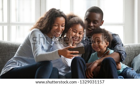 Happy young mom hold smartphone watch funny cartoons with mixed race family, relax together on sofa at home, smiling black parents have fun with little kids, enjoy movie or video on cellphone