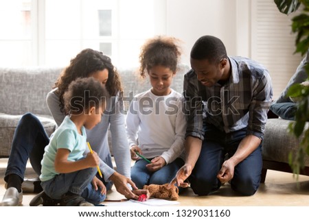 Young African American family spend time at home together drawing with colorful pencils, mixed race parents play with little kids painting picture, playing with toys sitting on floor in living room