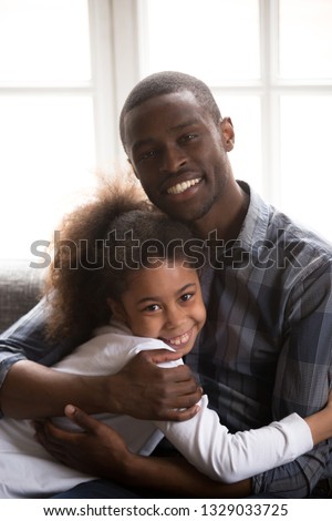 Portrait of young African American dad sit on couch hug teenage girl looking at camera, family picture of black smiling father hold in arms embrace little teen daughter relaxing on sofa at home