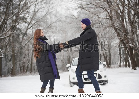 Holding by hands. Gorgeous young couple dancing in front of their white automobile in snowy forest.