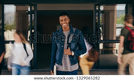 Portrait of african teenage boy at college campus with students walking by in motion blur. Happy young guy standing with his backpack looking at camera and smiling.