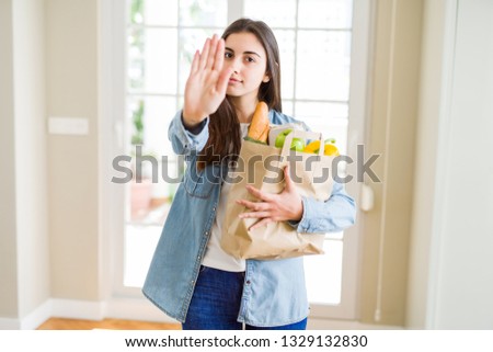 Beautiful young woman holding paper bag full of healthy groceries with open hand doing stop sign with serious and confident expression, defense gesture