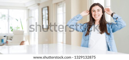 Wide angle picture of beautiful young woman sitting on white table at home Smiling pulling ears with fingers, funny gesture. Audition problem