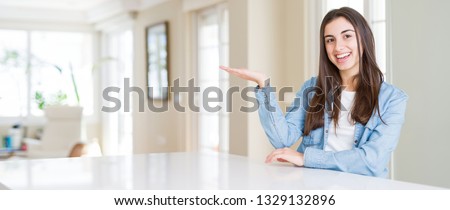 Wide angle picture of beautiful young woman sitting on white table at home smiling cheerful presenting and pointing with palm of hand looking at the camera.