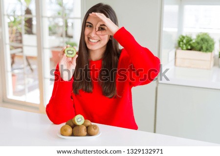 Beautiful young woman eating half fresh green kiwi with happy face smiling doing ok sign with hand on eye looking through fingers