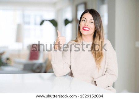 Young beautiful woman wearing winter sweater at home doing happy thumbs up gesture with hand. Approving expression looking at the camera showing success.