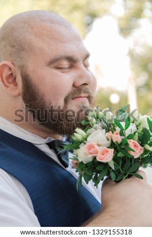 Stylish bearded fat groom with wedding bouquet in the park.