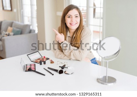Beautiful young woman using make up cosmetics cheerful with a smile of face pointing with hand and finger up to the side with happy and natural expression on face