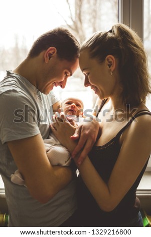 Woman and man holding on hands a newborn. On the background window. Mom, dad and baby. Portrait of young family. Happy family life. Man was born. Vertically framed shot. Royalty-Free Stock Photo #1329216800