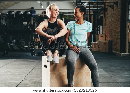 Two fit young female friends in sportswear laughing together while sitting on a box at the gym after a workout session Royalty-Free Stock Photo #1329219326