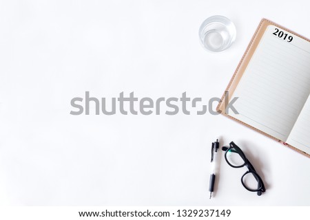 White office desk table with notebook, glass and pen. Top view with copy space, flat lay, 2019.