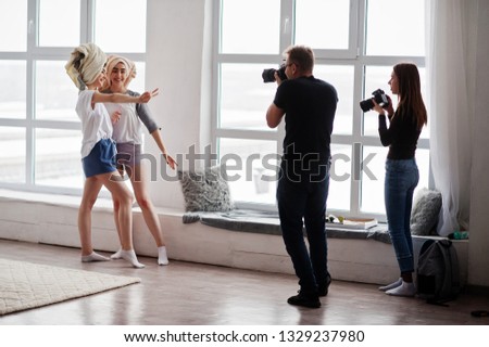 The team of two photographers shooting twins models girls on studio against large windows. Professional photographer on work.