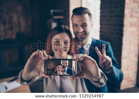 Close up photo she her business lady he him his guy laugh laughter hands arms hold smart phone make take selfies show v-sign say hi crazy blurred backdrop stand office wearing formal wear suits
