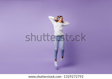 High-spirited lucky female student jumping in studio on isolated background. Full-length shot of girl in fashion white sweater and light blue jeans.