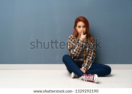 Young redhead woman sitting on floor showing a sign of silence gesture putting finger in mouth