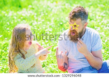 Dad and daughter sits on grass at grassplot, green background. Child and father posing with eyeglases photo booth attribute while speaking. Negotiation concept. Family spend leisure outdoors.