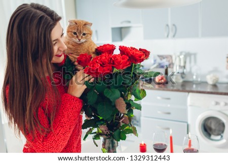 Young woman found red roses with candle, wine and gift box on kitchen. Happy girl smelling flowers with cat. Women's day Royalty-Free Stock Photo #1329312443