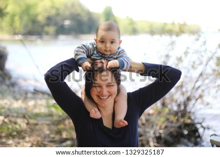 Young pretty mom and her little son having fun in front of spring forest fresh lake. Strong chubby baby is sitting on his mother neck. Woman is laughing. In soft focus, blur bokeh background.