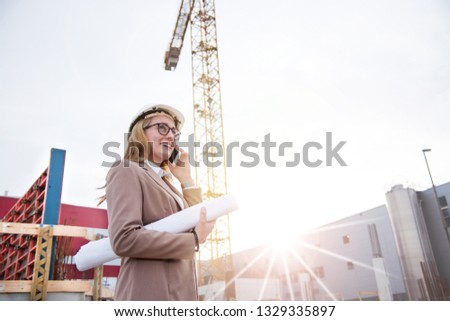 Woman as an architect with helmet, blueprint, plans phoning on a construction site with crane