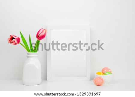Mock up white frame with Easter Eggs and tulip flowers on a shelf or desk. Easter concept. White color scheme. Portrait frame orientation.