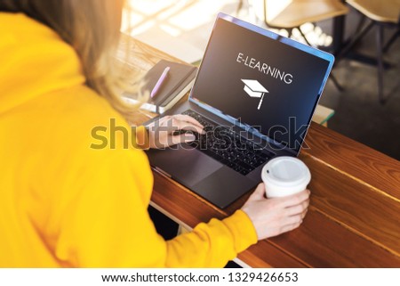 Close-up view of laptop screen with inscription e-learning. Sunny day. Girl in yellow hoodie sits in cafe at table,works on computer. Student doing homework. Business online education for adult.