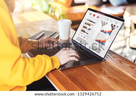 Close-up view of laptop screen with graphs, schedule, diagrams on monitor. Sunny day. Girl in yellow hoodie sits in cafe at table,works on computer. Freelancer works remotely.Business education online
