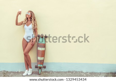 Beautiful blonde girl with long hair standing yellow wall in summer city. White body swimsuit board longboard. Free space for text. Selfie photo online call, social networks online, happy smiling.