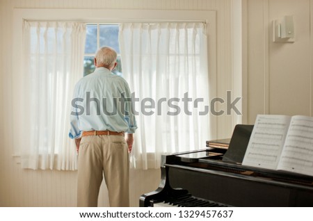 Senior man looking out the window next to a piano.