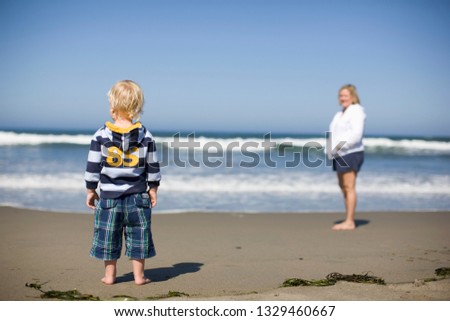 Young toddler standing on a sandy beach with his mother.