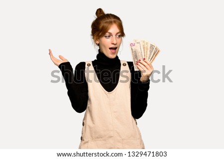 Young redhead woman taking a lot of money over grey background