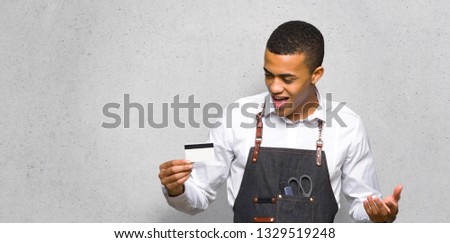 Young afro american barber man holding a credit card and surprised on textured wall
