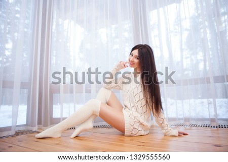 Portrait of beautiful happy stylish woman sitting on the floor near the window at the house. Winter forest at the window. Fashion woman lifestyle. Woman style, trendy outfit. Winter vacation.