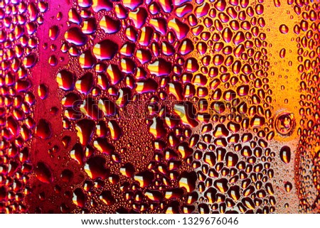 A close-up of droplets of water, rain or condensation on a sheet of transparent material or container beyond can be seen bright coloured neon colours such as a wet glass left to form condensation

